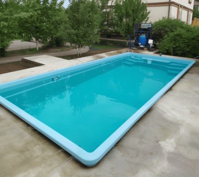 Голубой прямоугольный бассейн с бетонной площадкой и зелеными растениями